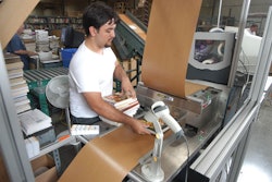 As the top and bottom web materials feed into the system, a packaging operator places a book on the bottom rigid board web.