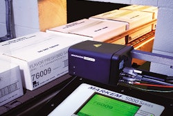 As taped shipping cases are conveyed through an opening in a wall, a coder prints the side of the box.