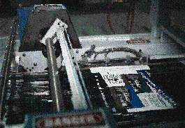 A plastic shutter enters the shrink wrapper, passing beneath the forming plow that wraps the film around and underneath. At the