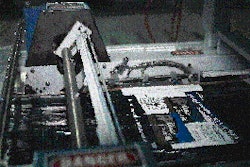 A plastic shutter enters the shrink wrapper, passing beneath the forming plow that wraps the film around and underneath. At the