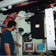 Video monitors in the background display an image of the printed film taken by a camera mounted above the press. Press operator