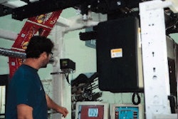 Video monitors in the background display an image of the printed film taken by a camera mounted above the press. Press operator