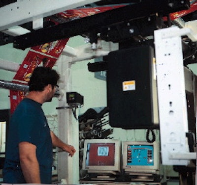 Video monitors in the background display an image of the printed film taken by a camera mounted above the press. Press operator