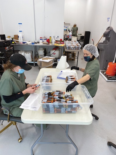 Manual label application is completed for a batch of pre-rolls bound for J-tube two-packs. The J-tubes are shrink-wrapped with printed film prior to labeling.