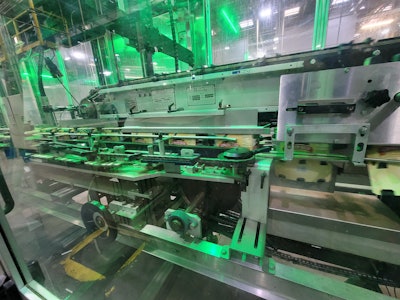 The 2D flat printed paperboard cartons are folded by a plow on the Marksman 750HS to enclose six packs on four sides, locking mechanically without glue.