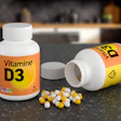Two white Vitamin D3 supplement bottles with yellow and white capsules spilled on dark surface