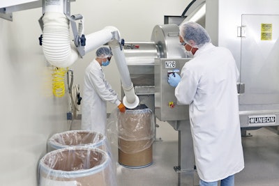 The blended batch is discharged into lined fiber drums, as dust is collected by a vacuum. Operator adjusts the mixer to rotate slowly during discharge.