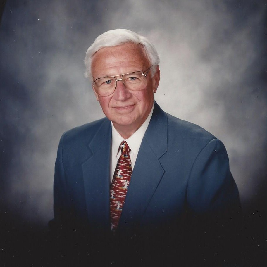 Professional portrait of an elderly man in navy blazer and patterned tie against neutral background