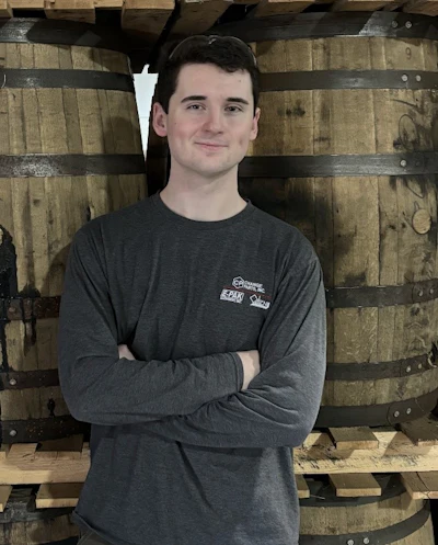 Young man in gray branded shirt posing with arms crossed against wooden barrel background