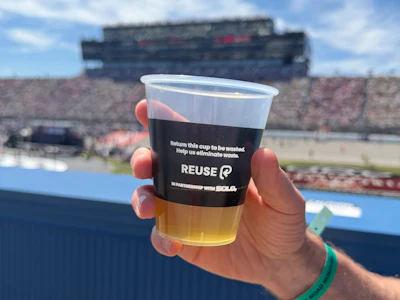 Reusable beverage cup with 'REUSE' branding held at a sports stadium with scoreboard visible