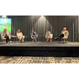 Five professionals participate in a panel discussion on stage with gray curtains and a presentation screen.