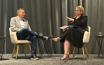 Two professionals engaged in a stage discussion with microphones and papers during a conference or interview event.