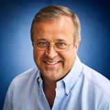 Professional headshot of a smiling man wearing glasses and a light blue button-up shirt against a blue gradient background