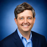 Professional headshot of a smiling man in navy blazer and light blue shirt against blue background