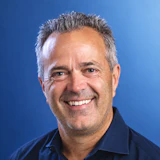Professional headshot of smiling middle-aged man in dark blue shirt against blue background