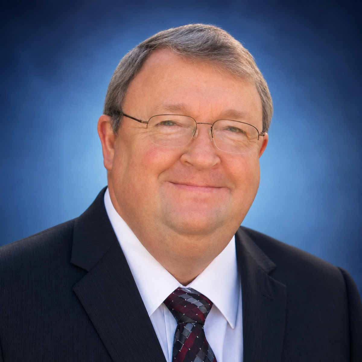 Professional headshot of a middle-aged man wearing glasses, dark suit, and burgundy tie against blue background