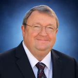 Professional headshot of a middle-aged man wearing glasses, dark suit, and burgundy tie against blue background