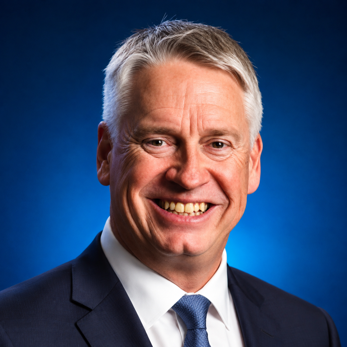 Professional headshot of a smiling man in dark suit and blue tie against blue background