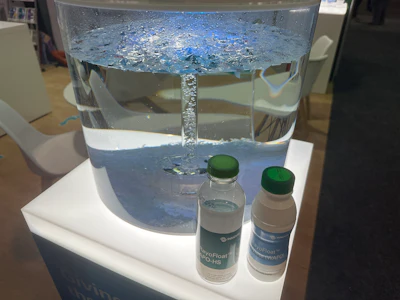A demonstration at Innovia's booth shows shredded PET and label material separating by density in water — recycled bottle flakes sink, the lighter polyolefin sleeve material floats to the top. RayoFloat APO-HS (clear) and White (WAPO) sample bottles sit in front of the tank.
