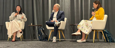 Three panelists seated in white chairs during a discussion event with gray curtain backdrop