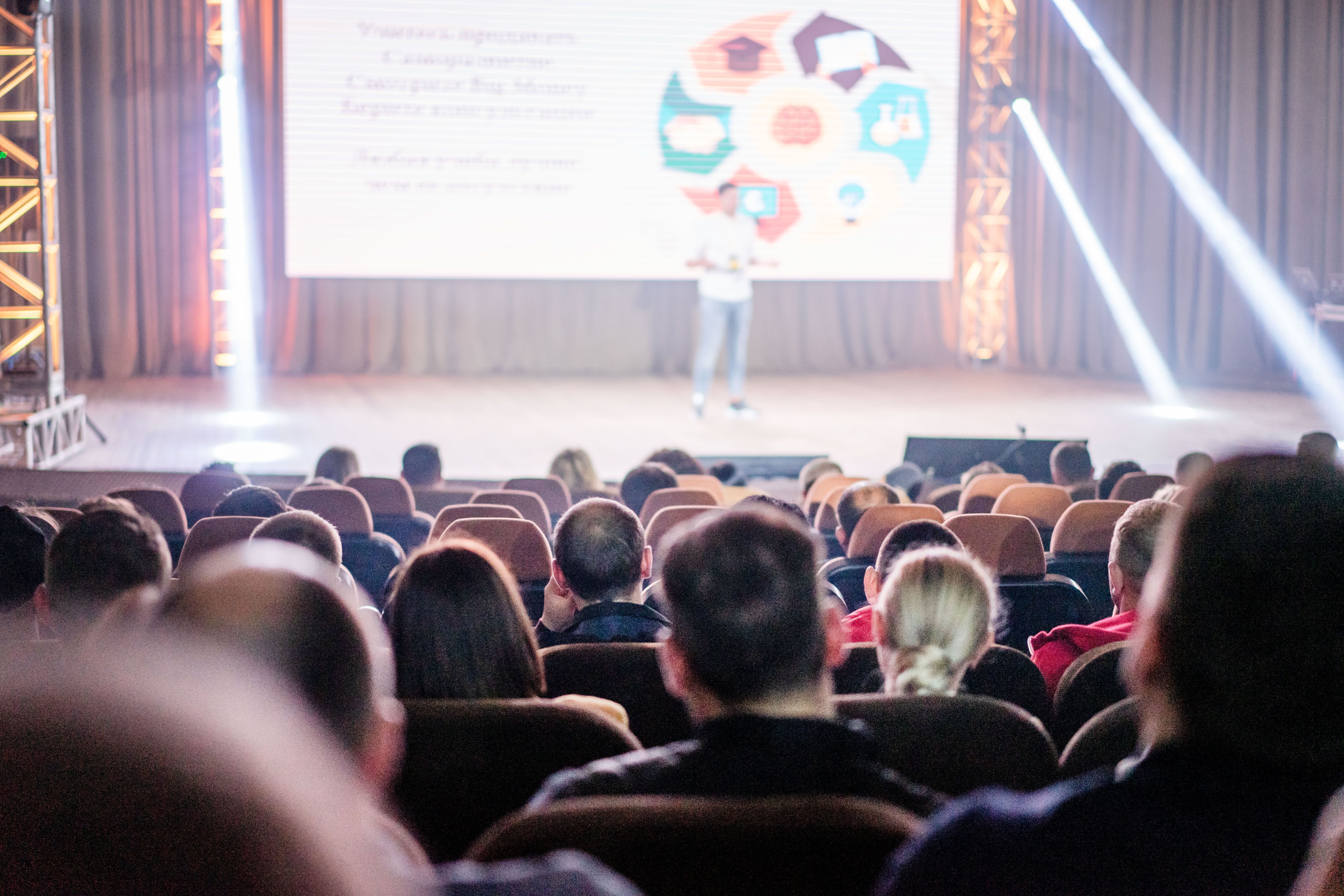 Audience seated in auditorium watching presenter deliver conference talk with colorful geometric shapes displayed on screen