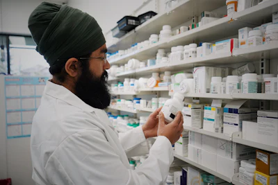 Pharmacist in white coat examining medication bottle in front of pharmacy shelves stocked with pharmaceutical products