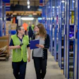 Two warehouse workers discussing inventory in a blue-shelved distribution center aisle