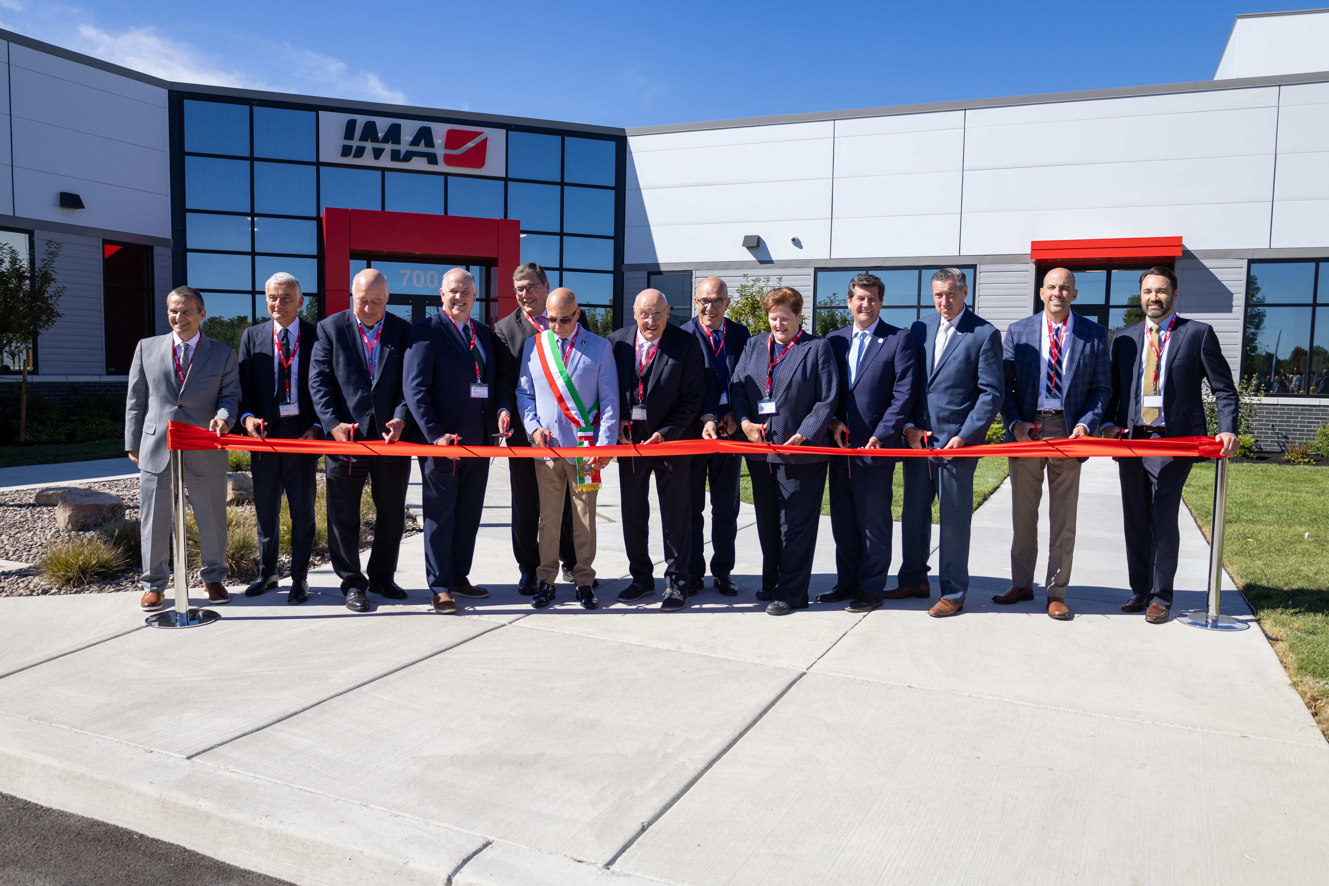 Group of business officials in suits cutting red ribbon at IMA facility ribbon-cutting ceremony