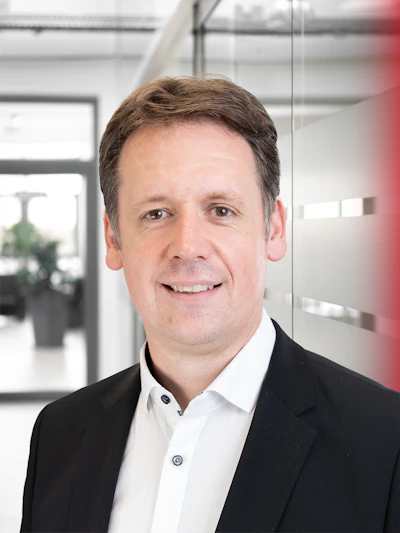 Professional headshot of a man in business attire smiling in a modern office environment