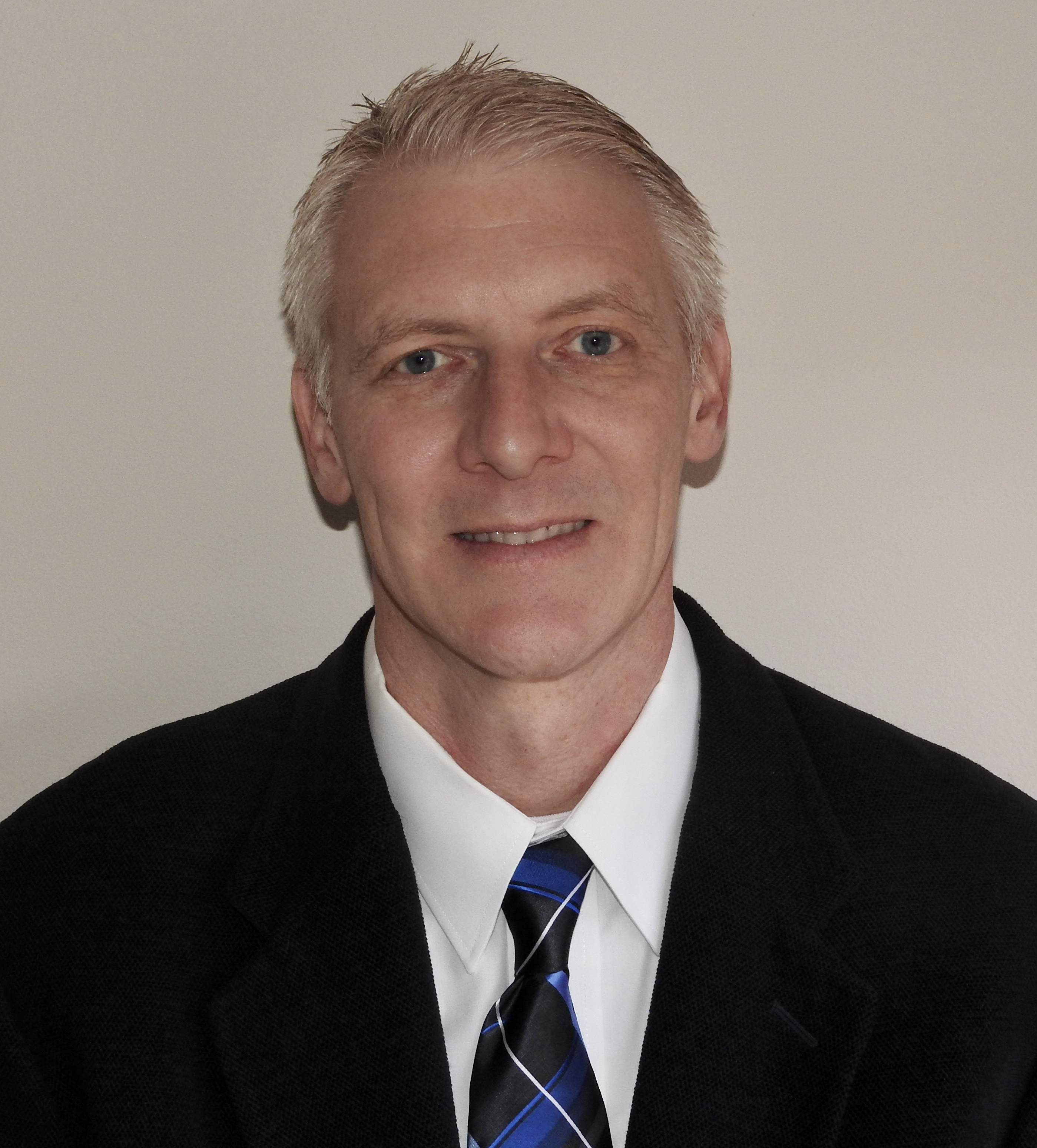 Professional headshot of a man in black blazer and blue striped tie against neutral background
