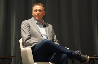 Man in gray blazer and jeans speaking at a professional event with microphone
