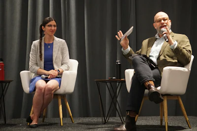 Two professionals engage in a panel discussion on stage, seated in white chairs with microphone and dark curtain backdrop.