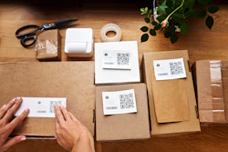 Overhead view of shipping workspace with kraft boxes labeled with QR codes, packing supplies, and green plant on wooden table