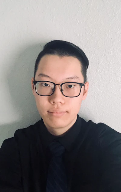 Professional headshot of a person wearing black-framed glasses and dark clothing against a white wall