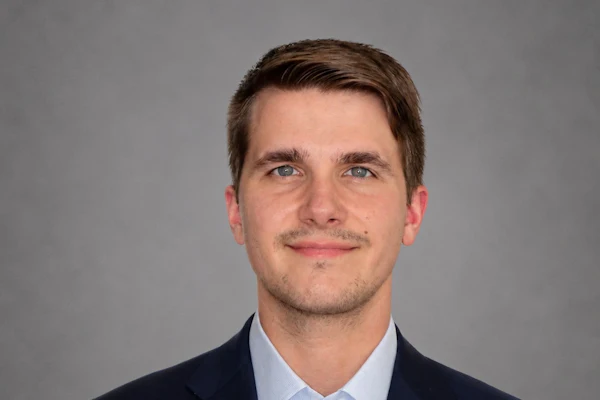 Professional headshot of a man wearing a dark blazer and light blue dress shirt against a gray background