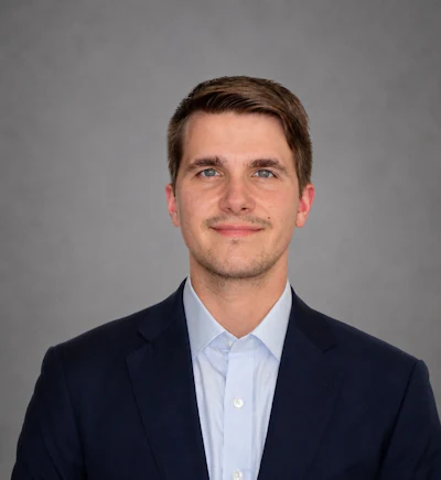 Professional headshot of a man wearing a dark blazer and light blue dress shirt against a gray background