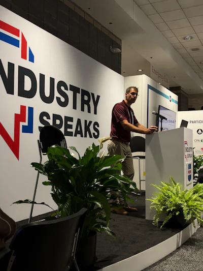 Speaker presenting at Industry Speaks event podium with branded signage and decorative plants
