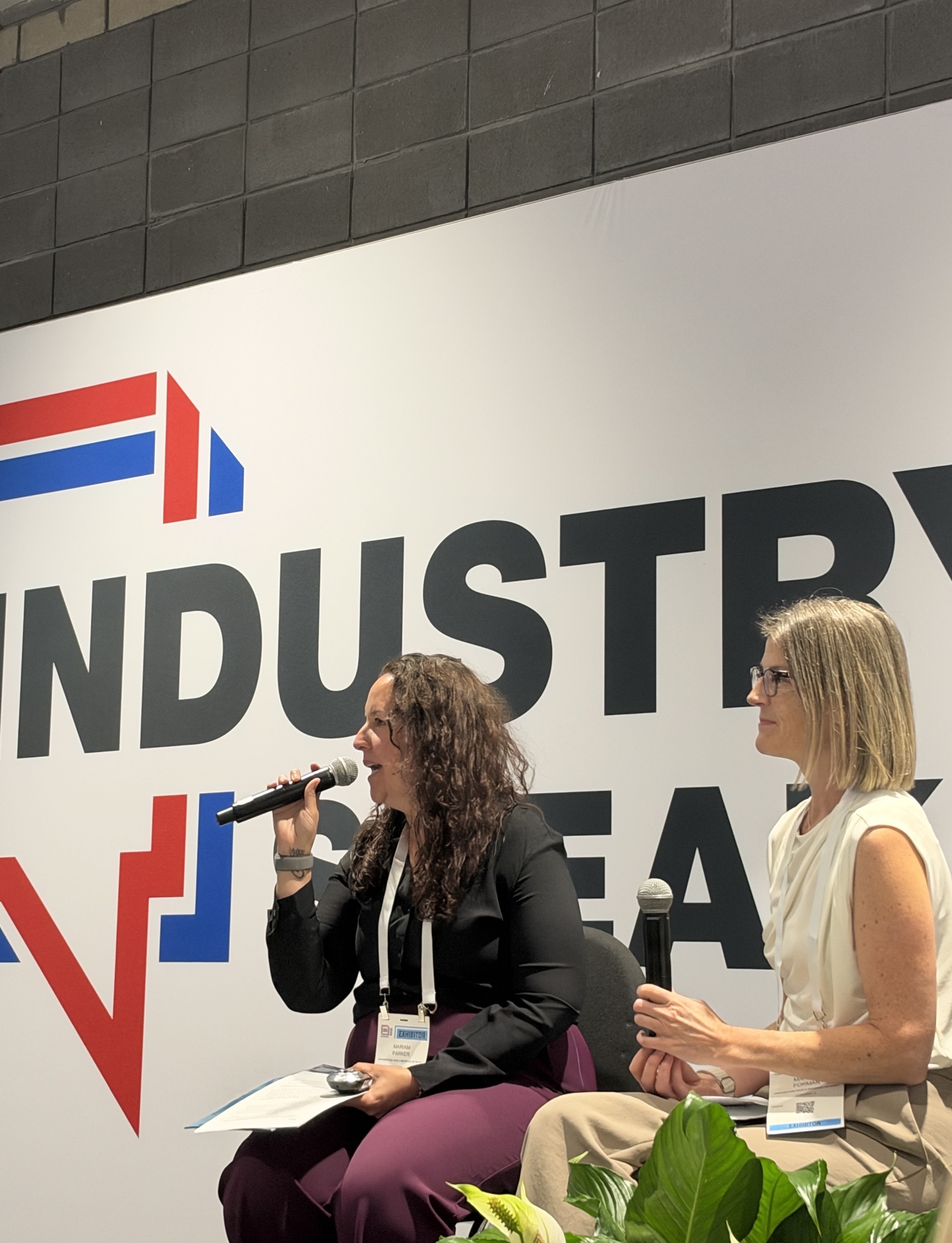 Two women speaking at a professional panel event with an industrial company logo displayed on the backdrop behind them.