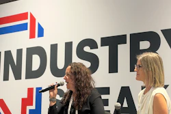 Two women speaking at a professional panel event with an industrial company logo displayed on the backdrop behind them.