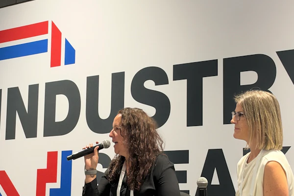 Two women speaking at a professional panel event with an industrial company logo displayed on the backdrop behind them.