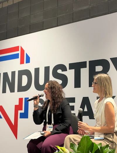 Two women speaking at a professional panel event with an industrial company logo displayed on the backdrop behind them.