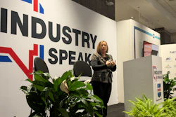 Professional woman speaking at Industry Speak conference with branded wall signage and decorative plants