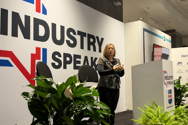 Professional woman speaking at Industry Speak conference with branded wall signage and decorative plants