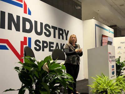 Professional woman speaking at Industry Speak conference with branded wall signage and decorative plants