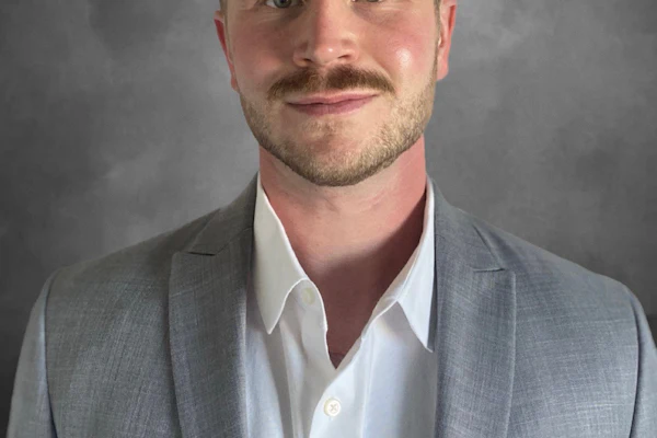 Professional headshot of a man in gray blazer and white shirt against neutral background
