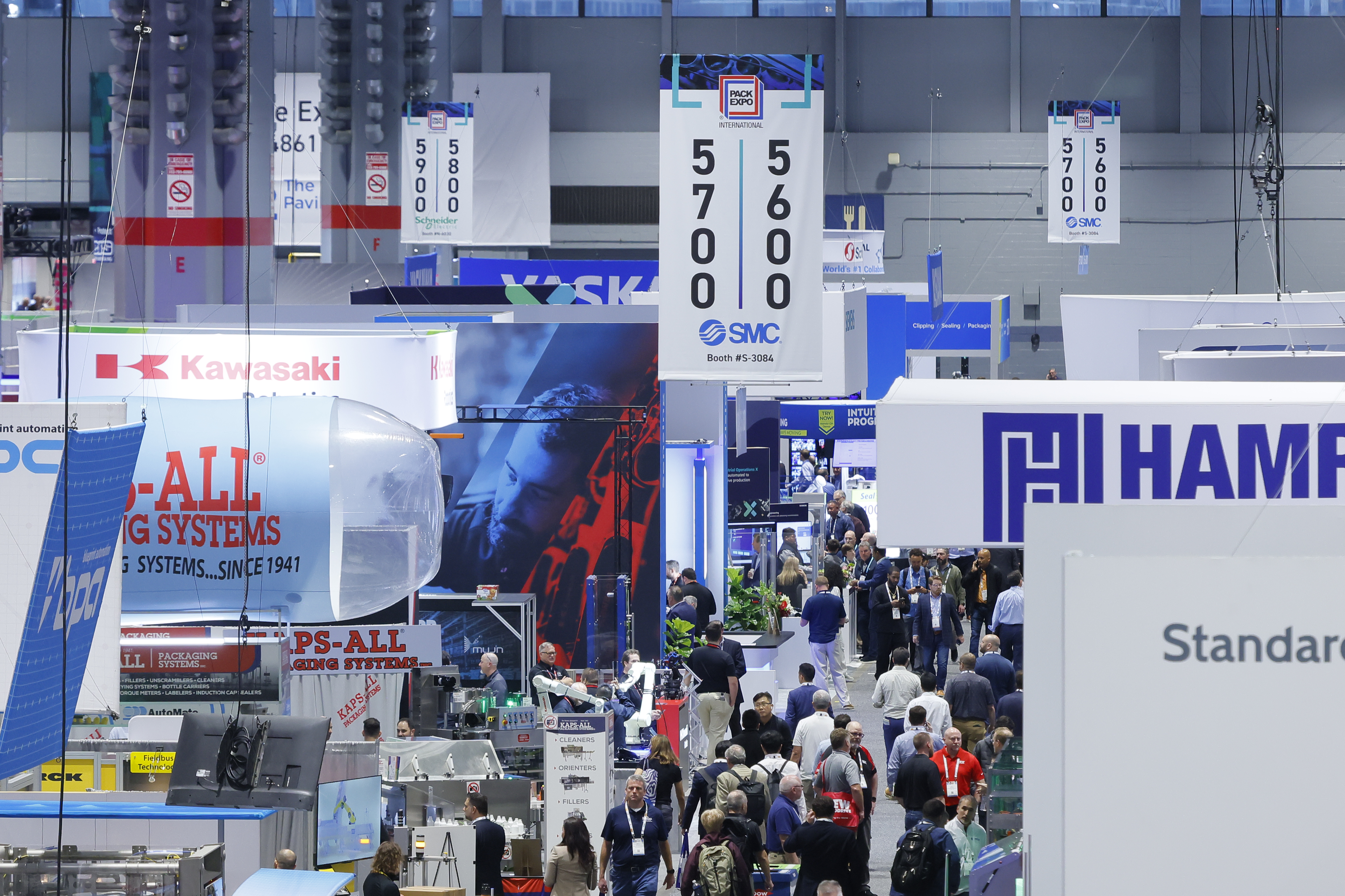 Crowded industrial trade show floor with multiple company booths displaying blue and red branded signage and digital displays
