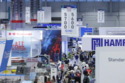 Crowded industrial trade show floor with multiple company booths displaying blue and red branded signage and digital displays