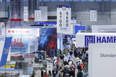 Crowded industrial trade show floor with multiple company booths displaying blue and red branded signage and digital displays