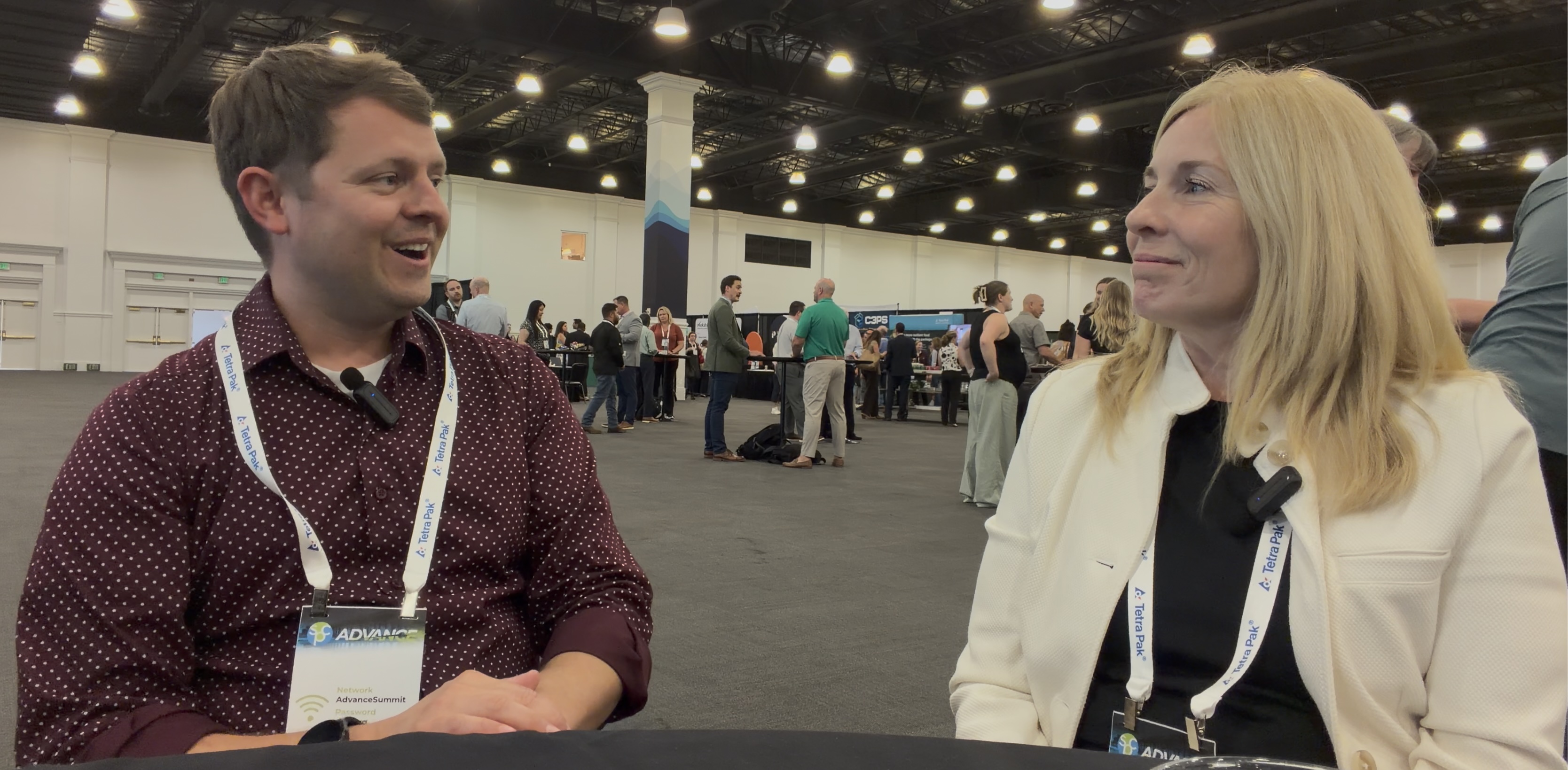 Two professionals wearing conference badges engage in conversation at an indoor event venue.