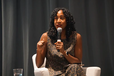 Woman speaking at a public event holding a microphone while seated on stage